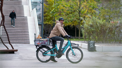 Si vede un uomo su una bicicletta da carico ibrida Cube Longtail blu che attraversa il centro città trasportando generi alimentari.