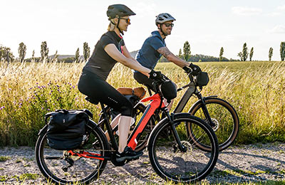 Si vede una coppia che percorre una strada sterrata con delle biciclette elettriche. Sullo sfondo si vede dell'erba alta.