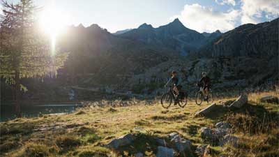Una coppia percorre un sentiero con le loro biciclette da trekking. Sullo sfondo si vedono le montagne dietro le quali spunta il sole.
