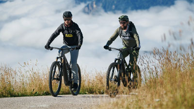 Due ciclisti pedalano sulle loro e-mountain bike su una pista ciclabile. Sullo sfondo si vedono le montagne.