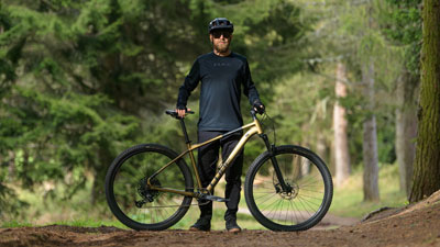Si vede un uomo dietro la sua mountain bike dorata, su un sentiero nel bosco.