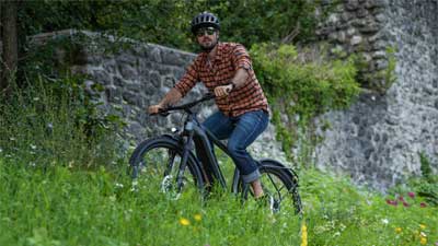 Un ciclista attraversa un prato con la sua mountain bike, sullo sfondo si vede un muro di pietra.