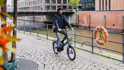 Un uomo va in bicicletta pieghevole blu, sullo sfondo ci sono case e un fiume.