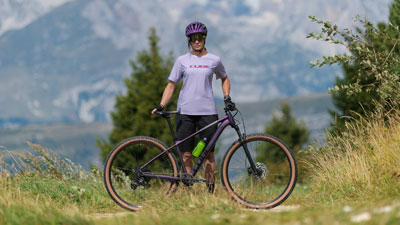 Si vede una donna in tenuta da ciclista viola, davanti a lei c'è una mountain bike viola. Sullo sfondo si vede un paesaggio montuoso.