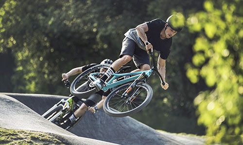 Ciclista salta con una moto da cross azzurra in un bike park, un altro ciclista con una bicicletta verde sullo sfondo.