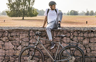 Si vede un uomo con un casco seduto su un muro. In primo piano c'è una bicicletta grigia e sullo sfondo si vede un paesaggio campestre.