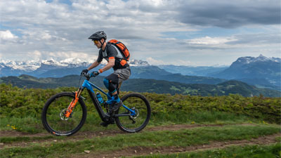 Si vede un uomo su una bicicletta elettrica ibrida Cube Stereo blu con dettagli arancioni. Sta pedalando su un sentiero escursionistico e sullo sfondo si vedono le montagne.