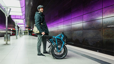 Si vede un uomo in una stazione della metropolitana, davanti a lui c'è una bicicletta pieghevole blu.