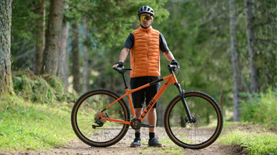 Un uomo è in piedi dietro alla sua mountain bike arancione Cube, su un sentiero nel bosco.
