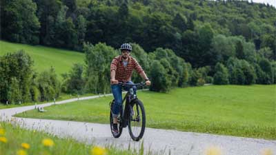 Un ciclista percorre una pista ciclabile asfaltata con la sua mountain bike, sullo sfondo si vede un paesaggio rurale.