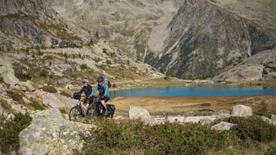 Una coppia percorre in bicicletta un paesaggio roccioso, sullo sfondo si vedono montagne e un lago.
