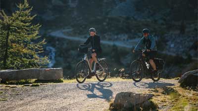 Una coppia va in mountain bike su una pista ciclabile, sullo sfondo si vede un paesaggio montuoso.