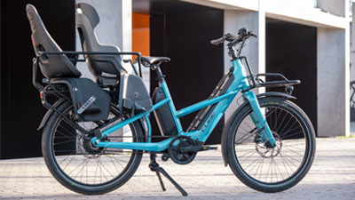 Si vede una bicicletta da carico ibrida Cube Longtail blu, con due seggiolini per bambini attrezzati.