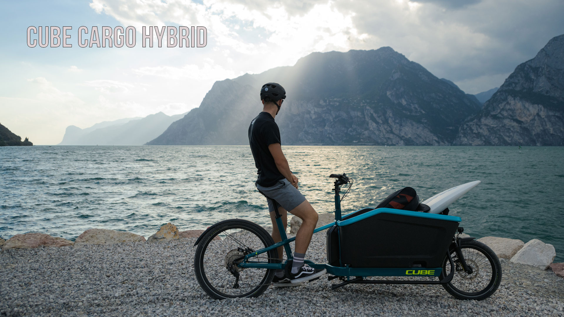 Si vede un uomo su una bicicletta da carico blu, in piedi sulla riva di un lago che osserva le montagne sullo sfondo. L'immagine è intitolata Cube Cargo Hybrid.