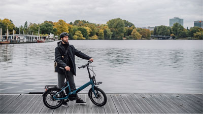 Un uomo è in piedi accanto alla sua bicicletta pieghevole blu su una passerella. Sullo sfondo si vedono l'acqua e una città.
