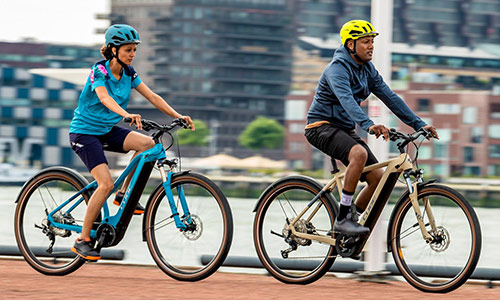 Un uomo e una donna stanno pedalando su una pista ciclabile, sullo sfondo si vede una grande città.