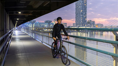 Si vede una donna in bicicletta che attraversa un sottopassaggio, sullo sfondo si intravede una grande città.