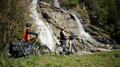 Una coppia spinge le biciclette attraverso un prato, sullo sfondo si vede una cascata.