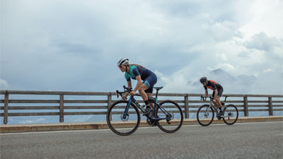 Si vedono due ciclisti su biciclette da corsa, sullo sfondo una staccionata di legno e nuvole.
