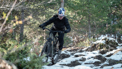 Una ciclista sta percorrendo con la sua mountain bike un sentiero boscoso e accidentato, in parte ancora coperto di neve.