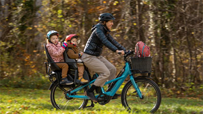 Si vede una madre su una bicicletta da carico Cube Longtail blu. Sul portapacchi sono seduti due bambini piccoli.